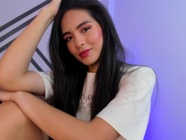 MadisonSmitt has long black hair, pink eyeshadow, red lipstick, and is wearing a white shirt. She's sitting with one arm on her knee, looking at the camera.