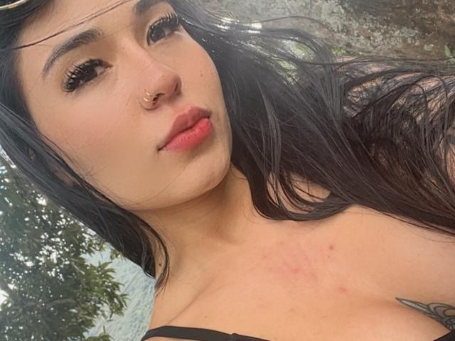 AlahiaRouse has long, black hair that flows to the right. She has a nose ring in her left nostril and full, pinkish-red lips. Her skin is light with some redness on her chest. She has dark, thick eyebrows and wears subtle makeup. A black strap is visible on her right shoulder. The image is a close-up with a slightly blurred, greenish background. She looks to the left with a calm expression. The lighting is soft, emphasizing her facial features.