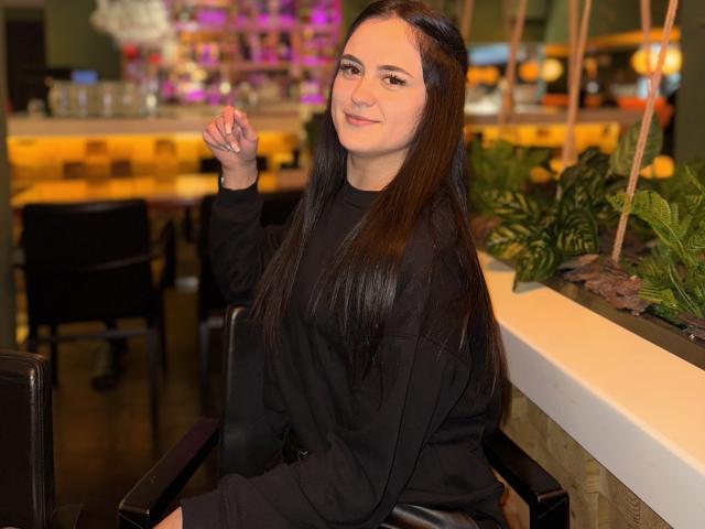 MarySweetSuprem with long brown hair, black shirt, sitting in a chair, raising her right hand, smiling, in a dimly lit restaurant with green plants and warm lighting.