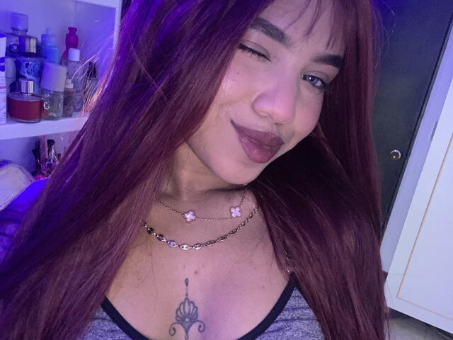 KathaThoms with long red hair, winking, and pouting with dark lipstick. She's wearing a necklace with pink flowers and a gray top, showing a tattoo on her tits.