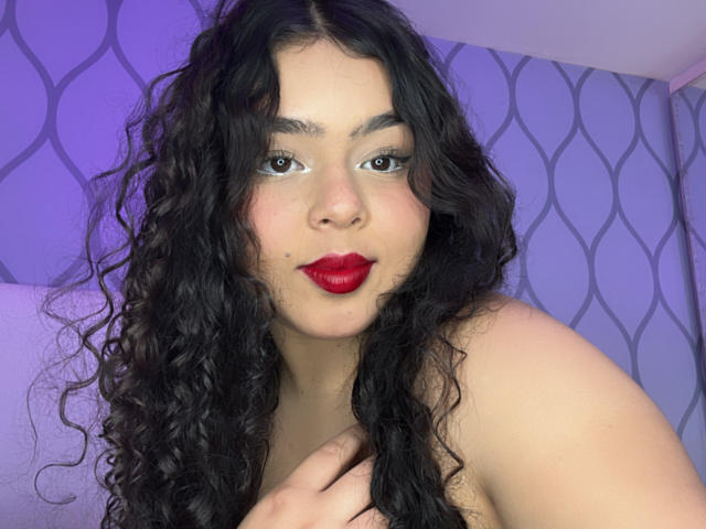 AprielKeith has curly black hair, red lipstick, and a soft smile. Her brown eyes are looking at the camera with a slight, sexy, confident expression. She's topless.