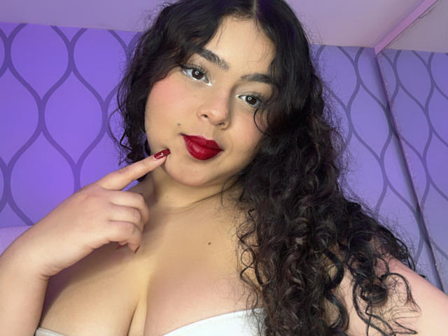 AprielKeith with curly black hair, red lipstick, and white strapless top, pointing at her red-painted lips, showing her large, round boobs. Her skin is light, and she's looking at the camera.