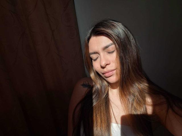 AmaiaSalvatoree with long brown hair, eyes closed, sunlight on face, subtle smile, white top, tattoo on right shoulder, brown curtain in background.