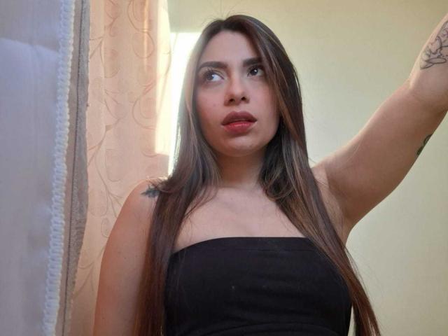 AmaiaSalvatoree has long brown hair, red lipstick, and is wearing a black strapless top. She's looking to the side with her arm raised. Visible tattoo on her arm.