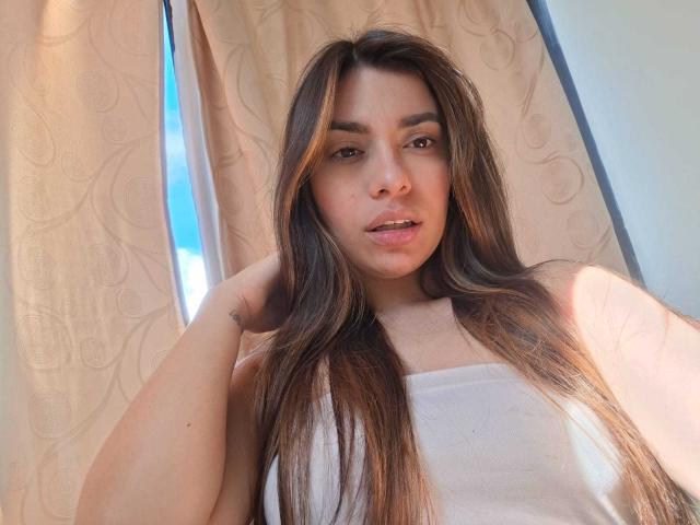 AmaiaSalvatoree with long brown hair, wearing a white tank top, has a relaxed expression, hand in hair, small tattoo on wrist, light skin, subtle makeup, natural light.