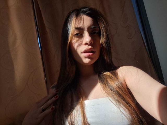 AmaiaSalvatoree with long brown hair, sunlit, wearing a white strapless top, hand with red nails, subtle smile, natural light, soft shadows, pinkish curtain in background.