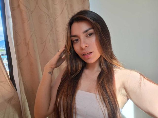 AmaiaSalvatoree with long brown hair, light skin, brown eyes, small tattoo on left hand, wearing a white strapless top, standing in front of beige curtains, smiling softly.