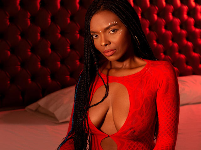 MonyWow with dark skin, long braids, wearing a red lace bodysuit with a deep cleavage, face decorated with small white dots, red tufted headboard in background.