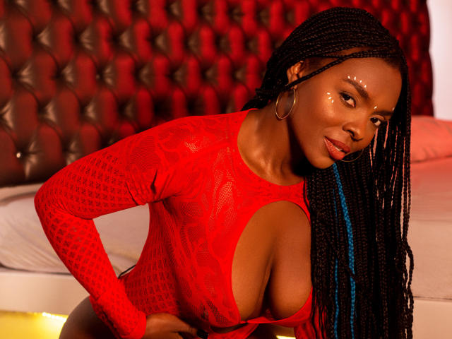 MonyWow with dark skin, long braids, wearing a red mesh lingerie dress, showing big boobs, large cutout, shiny face, gold hoop earrings, leaning forward, sexy smile, red background.