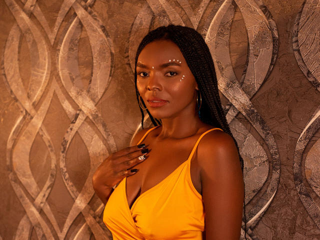 MonyWow with dark skin, braided hair, and sparkling face dots, wearing a yellow dress, hand on chest, black nails, hoop earrings, looking at camera, warm lighting.