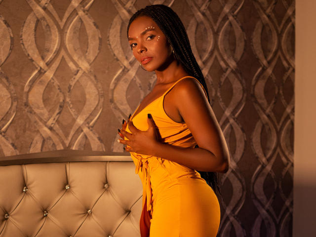 MonyWow in a yellow dress, covering her tits with one hand, standing by a tufted beige headboard, with braided hair and warm brown skin, looking to the side.