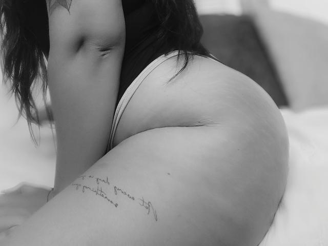 Black-and-white photo of AiuniHot's curvy ass in white panties, showing a tattoo on her right thigh, with long dark hair partially visible, kneeling on a bed.