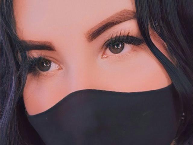 AiuniHot's face with dark, wavy hair, thick eyebrows, and green eyes, partially covered by a black mask, showing only eyes and forehead. She has long, thick, black eyelashes.