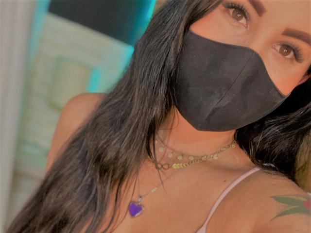 AiuniHot with long black hair, wearing a black mask, gold necklaces, purple heart pendant, and a green tattoo on the right shoulder, showing brown eyes.