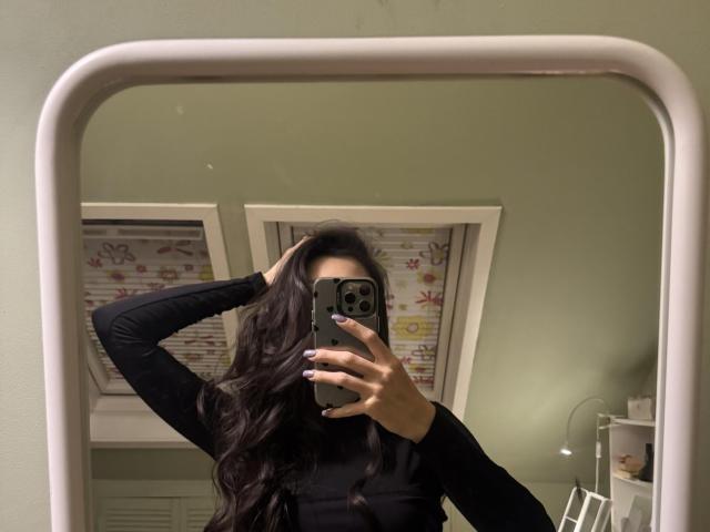 AnitaLiberty with long dark wavy hair, black long-sleeve shirt, holding phone in front of mirror, covering face, hand in hair, green wall, white-framed mirror, visible window.