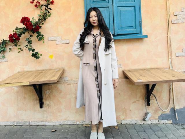 AnitaLiberty stands with long black hair, wearing a pale pink dress with black lines, white coat, and white shoes, against a peach wall with blue window.