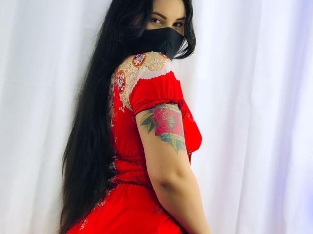 AiuniHot with long black hair, wearing a red lace dress, black mask, and a rose tattoo on her right arm, looking back over her shoulder.