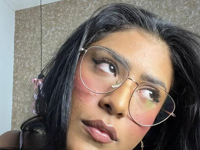 Babilett has dark brown skin, black hair, and wears round glasses with long eyelashes. Her pink cheeks and glossy lips are visible. She looks to the side with a pout.