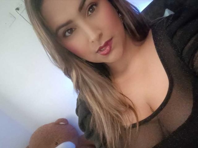 Marcee69 with long brown hair, wearing a sheer black top, showing cleavage, red lipstick, and a slight smile, holding a brown teddy bear in a soft-lit room.