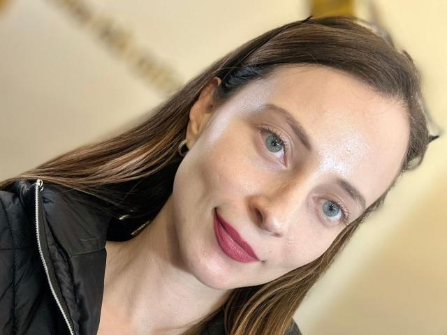 AmbeerWood has fair skin, blue eyes, and brown hair. She's wearing a black quilted jacket, with pink lipstick and a slight smile. Her face is shiny and she's looking at the camera.