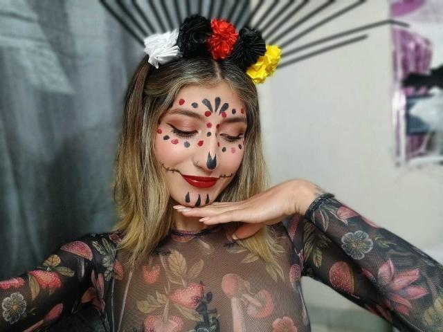 Kaiix met een beschilderd gezicht, rode stipjes, zwarte lijnen en een bloemenhoofddeksel, draagt een doorschijnende zwarte top met bloemen, rode lippenstift, blond haar, en raakt haar kin aan, op een kleurrijke, gedetailleerde foto..