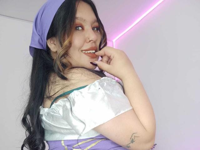 CaroHotcg with long black hair, purple headscarf, white off-shoulder top, purple dress, smiling, finger on lips, tattoo on right arm, pink neon light in background.