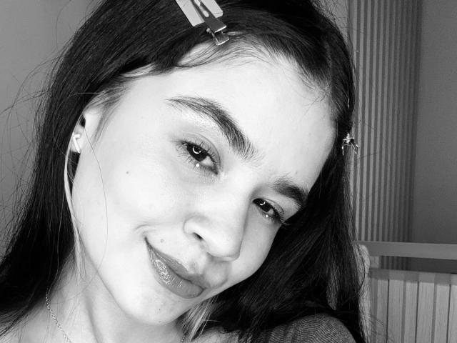 Selfie in bianco e nero di IsabellaMarynn con un sorriso carino, capelli scuri raccolti con delle clip, auricolari nelle orecchie, testa leggermente inclinata e una piccola lentiggine sulla guancia destra..