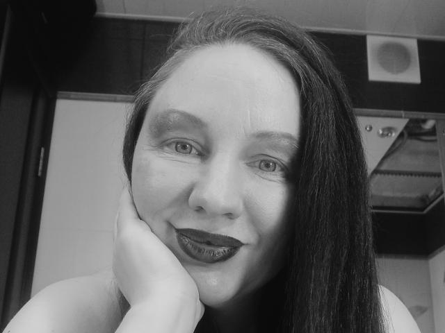 Black-and-white selfie of DashaHart with long dark hair, dark lipstick, and hand resting on cheek. She's smiling with subtle makeup, showing light skin and small eyes.