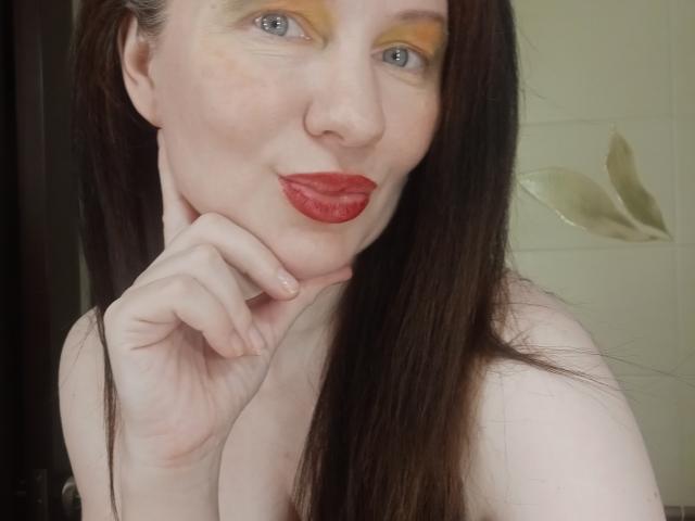 DashaHart with long brown hair, yellow eyeshadow, red lipstick, pale skin, blue eyes, and red-painted fingernails, smiling slightly, touching her chin. No clothes visible.