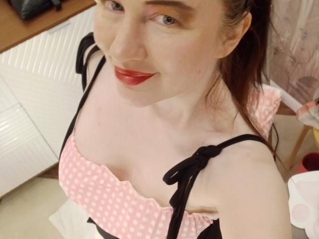 DashaHart with pale skin, red lipstick, and brown hair in a ponytail, wearing a pink polka dot top with black straps, showing her tits. She's smiling at the camera.