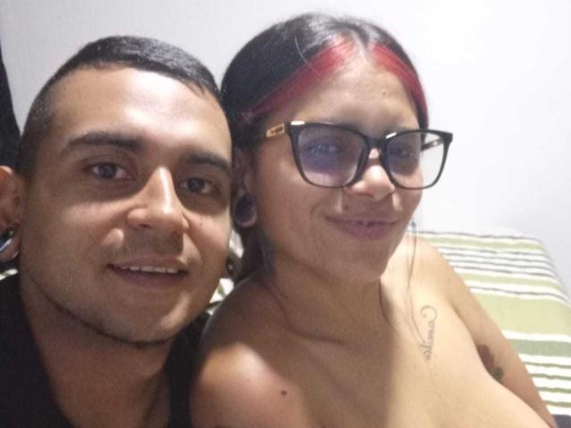 ElliiandCarl69 has short black hair and a slight beard, smiling next to a topless woman with red highlights, glasses, and a tattoo on her chest, showing her tits.
