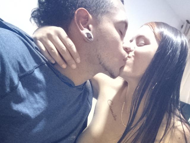 Elliiand69 with brown hair, small tits, kissing Carl69, who has dark hair, ear gauges, and a tattoo on his chest, wearing a blue shirt. Both are topless, eyes closed.