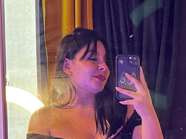 EmaJames macht ein Selfie vor einem Spiegel mit einem lila Handy und zeigt ihre nackten Schultern und ihr schwarzes Kleid. Ihre Brüste sind leicht sichtbar, und sie lächelt leicht..