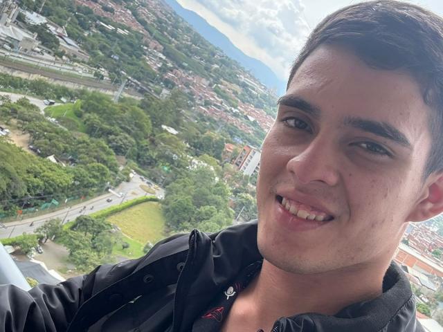 KainNoir has short brown hair, a cute smile, and a black jacket, with a slightly visible pink shirt. He's smiling at the camera, showing his teeth.