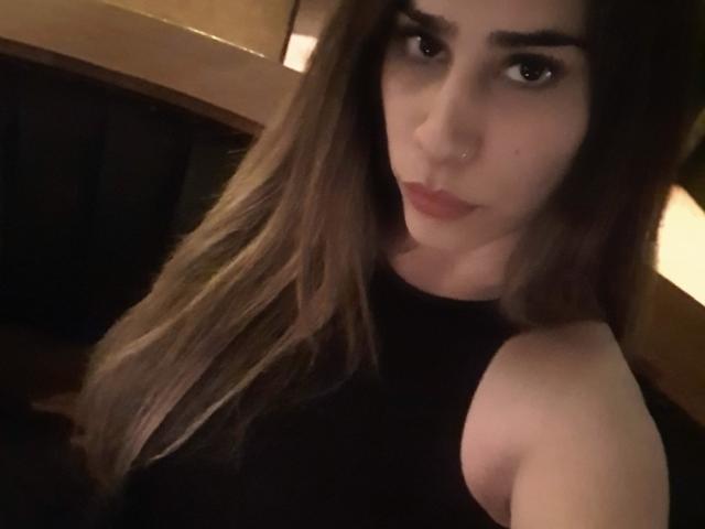 NahiaraBiel has long brown hair, dark eyebrows, and is wearing a black sleeveless top, showing her bare shoulders and looking at the camera with a slight pout.