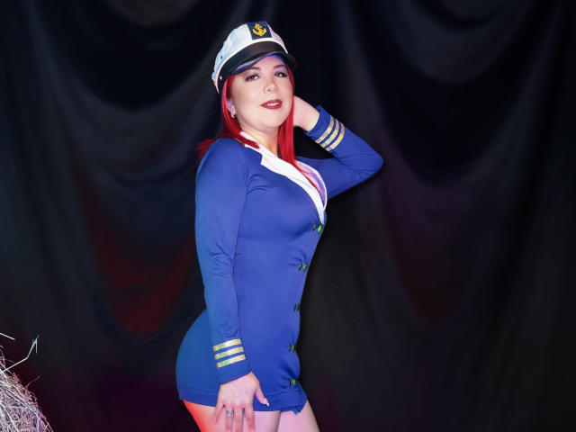 CorineRay in a blue pilot outfit with a white cap, hand on her head, showing her tits and ass, red lipstick, and red hair, against a dark background.