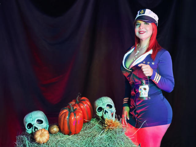 CorineRay with red hair, wearing a navy and white sailor outfit, holding a mug, standing next to a hay pile with skulls, pumpkins, and a small gourd.