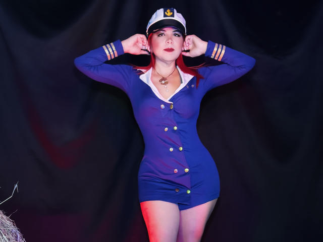 CorineRay in a tight blue pilot dress with gold buttons, white collar, and cap, standing with hands in hat, red lipstick, white skin, and red hair. Her tits are small.