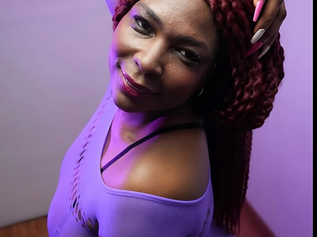 Ebonyy69 with red braided hair, smiling, wearing a purple off-shoulder top, showing her shoulder and black strap, under purple lighting, with hand on head.