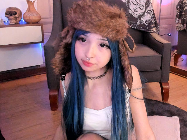 MillaHottLove69 with blue hair, wearing a furry hat, white dress, black choker, and nose ring, sitting on a rug, showing her tits slightly, in a cozy room.