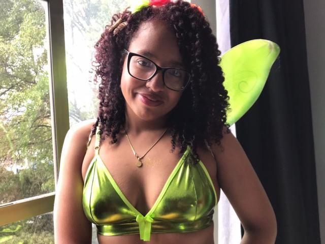 ArtemisaS with curly dark hair, wearing green fairy wings and a shiny green bikini top, showing her tits, smiling at the camera, wearing glasses, and a necklace.