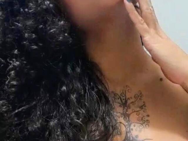 RosannaBlue's curly black hair, a hand on her neck, and a tree tattoo on her chest. Her skin is tan. Her tits are slightly visible.