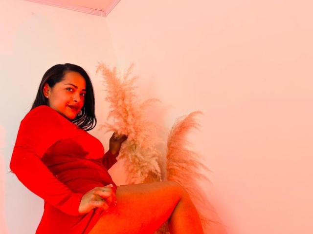DannaRamiirez in a red dress, sitting with one leg up, hand on thigh, puffy pinkish-white floss near her, looking at camera with red lips, dark hair, slight smile.
