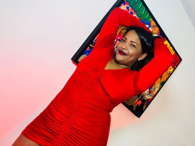 DannaRamiirez in a tight red dress with ripples, arms raised, smiling, showing her tits, standing in front of a colorful painting, white wall, red light.
