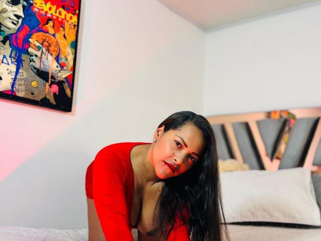 DannaRamiirez with long black hair, red top, and visible cleavage, leaning forward on a bed, looking at the camera, in a brightly lit room with colorful wall art.