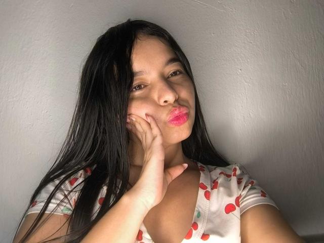 LissyCarter with long black hair, puffy pink lips, hand on cheek, wearing a white shirt with red cherry patterns, against a grey wall, looking slightly to the side.