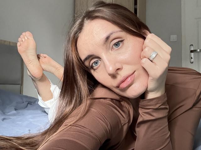 NastyGalq with long brown hair, blue eyes, and a brown long-sleeve shirt, laying on a bed, head on hand, showing a ring, with feet up.