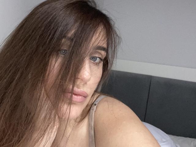 NastyGalq with long brown hair covering her eyes, grey eyes, and pink lips, wearing a beige tank top, looking at the camera with a neutral expression.
