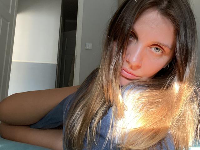 NastyGalq has long brown hair, blue eyes, and is lying down with sunlight highlighting her face. She's wearing a blue shirt, looking seductively at the camera.