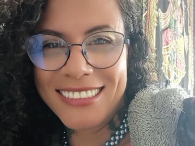 RosannaBlue's face with curly black hair, wearing glasses and a grey jacket with a furry collar, smiling with white teeth and pink lips. Her eyes are green.
